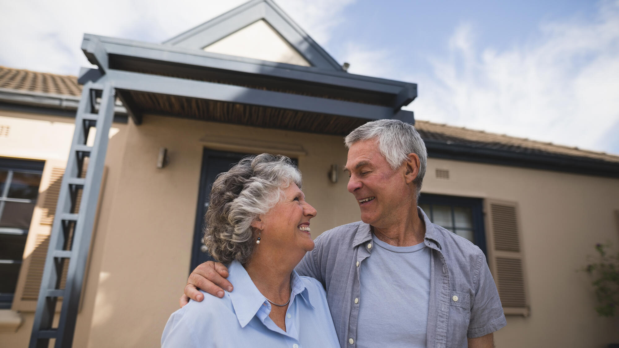 An elder couple standing outside of their home, smiling at one another